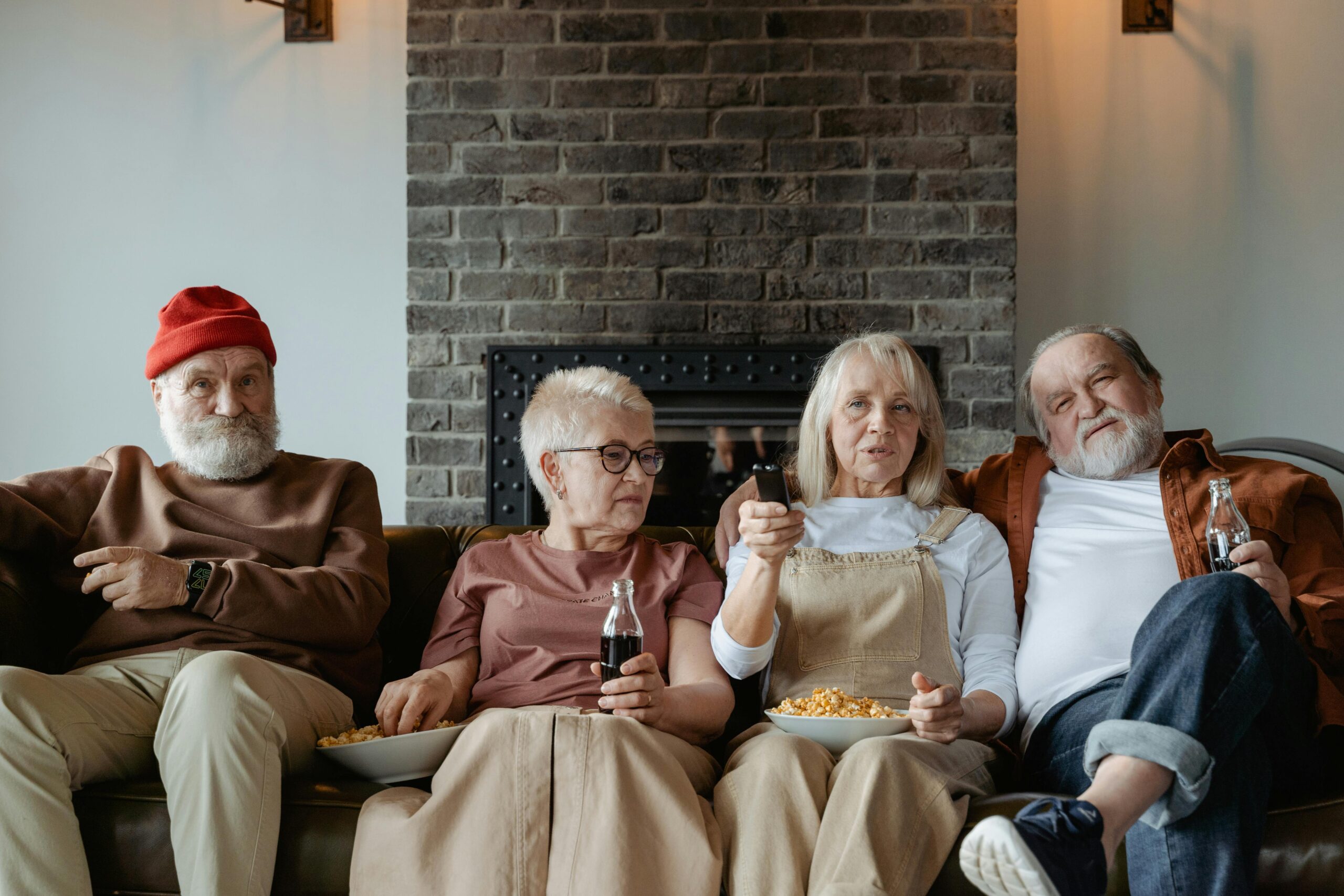 Four elderly friends enjoying a cozy TV time indoors with snacks and drinks.