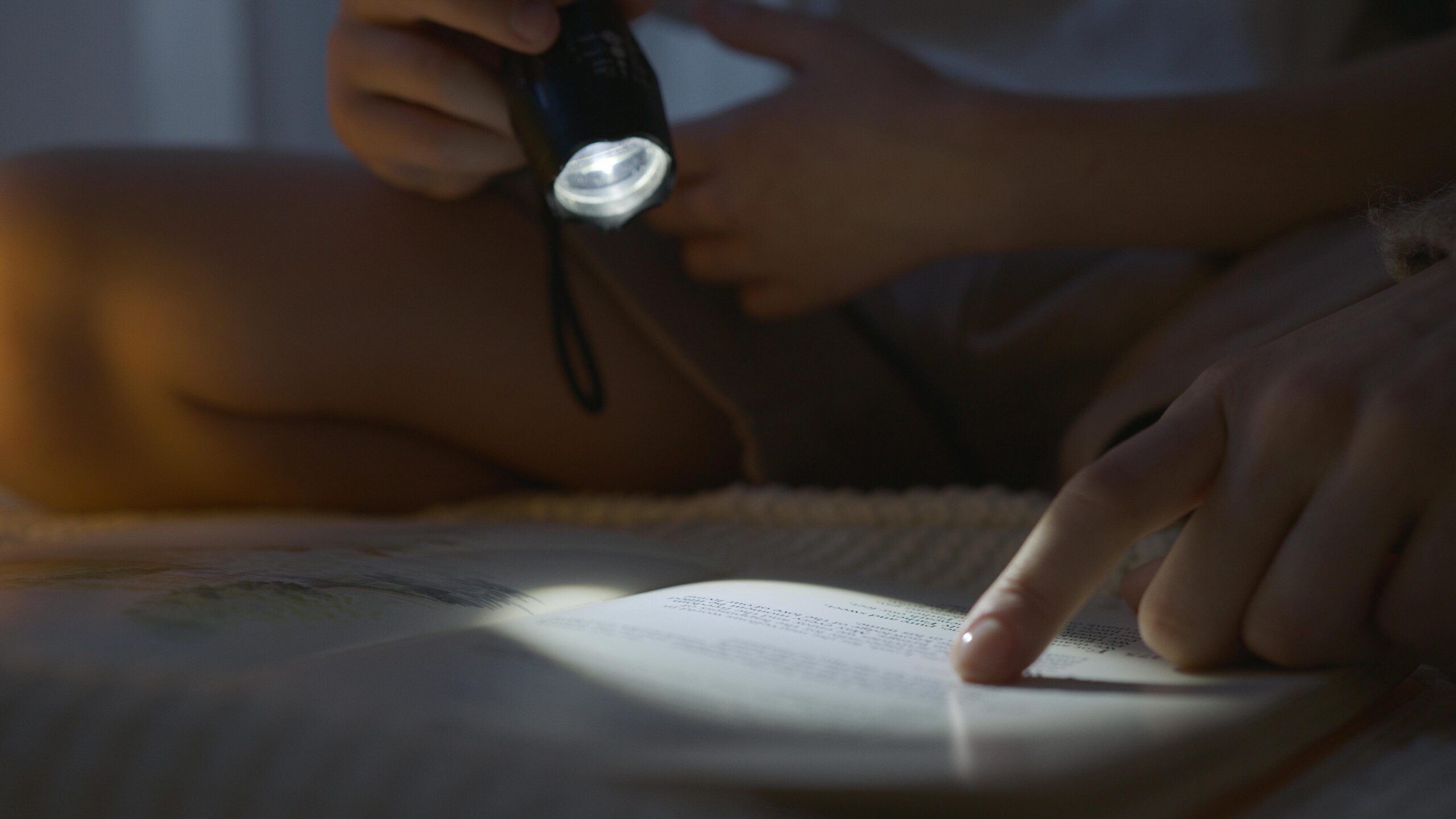 A person reads a book in dim light using a flashlight, focusing on the text with a finger.