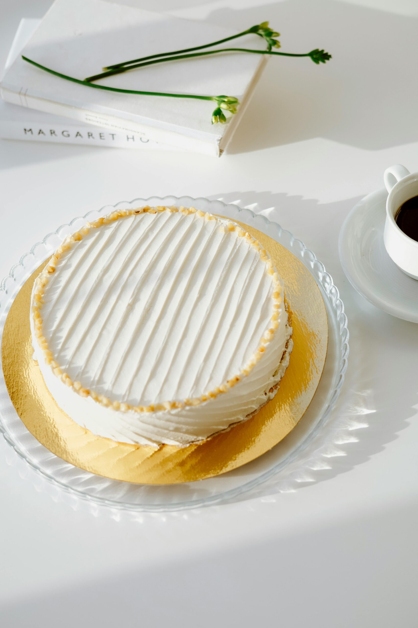 Minimalist white cake on a glass plate with a cup of coffee and books on a sunlit white table.